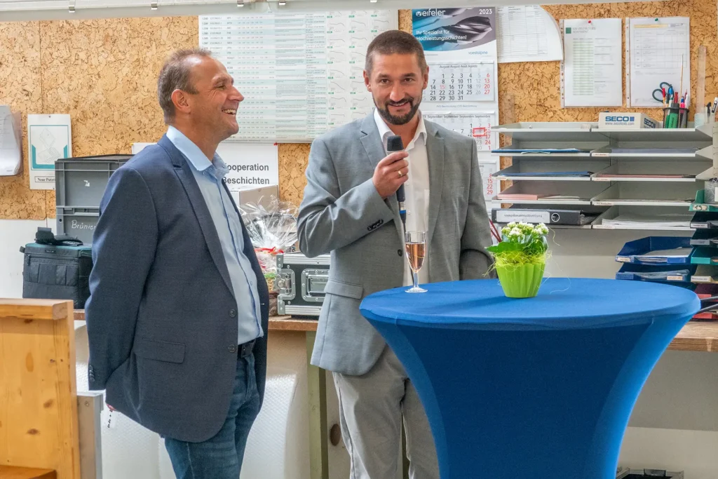 René Liebermann und Patrick Hammerschmidt stehen lächelnd an einem Tisch, Patrick Hammerschmidt hält Mikrofon in der Hand