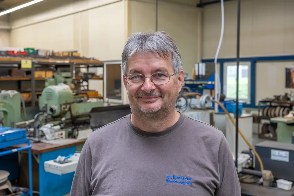 Portraitfoto Fredi Forkel: lächelnder Herr mit Brille und grau melierten Haaren in dunkelgrauem Technotrans-T-Shirt mit Maschinen im Hintergrund