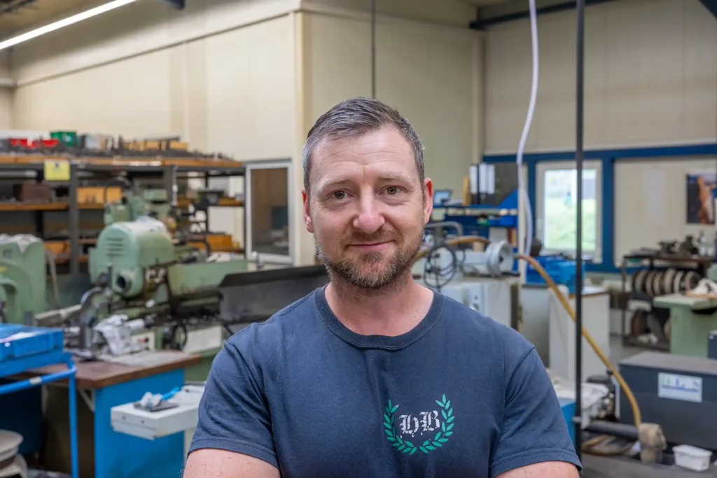 Portraitfoto Mario Seifferth: lächelnder Herr mit kurzen braunen Haaren, Vollbart und dunkelblauem T-Shirt mit Maschinen im Hintergrund